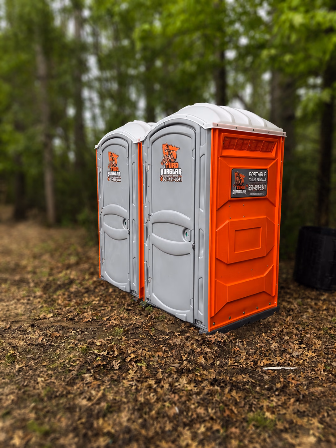 Two portable toilets, one gray and one orange, standing among fallen leaves in a wooded yard.