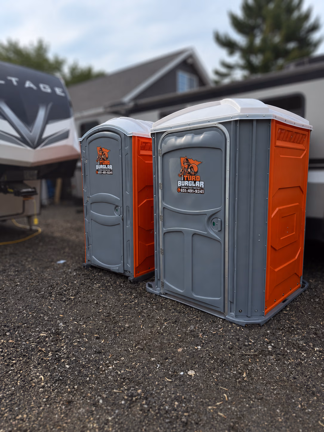A row of seven green portable toilets with white roofs standing on wet pavement in a park-like area with trees in the background.