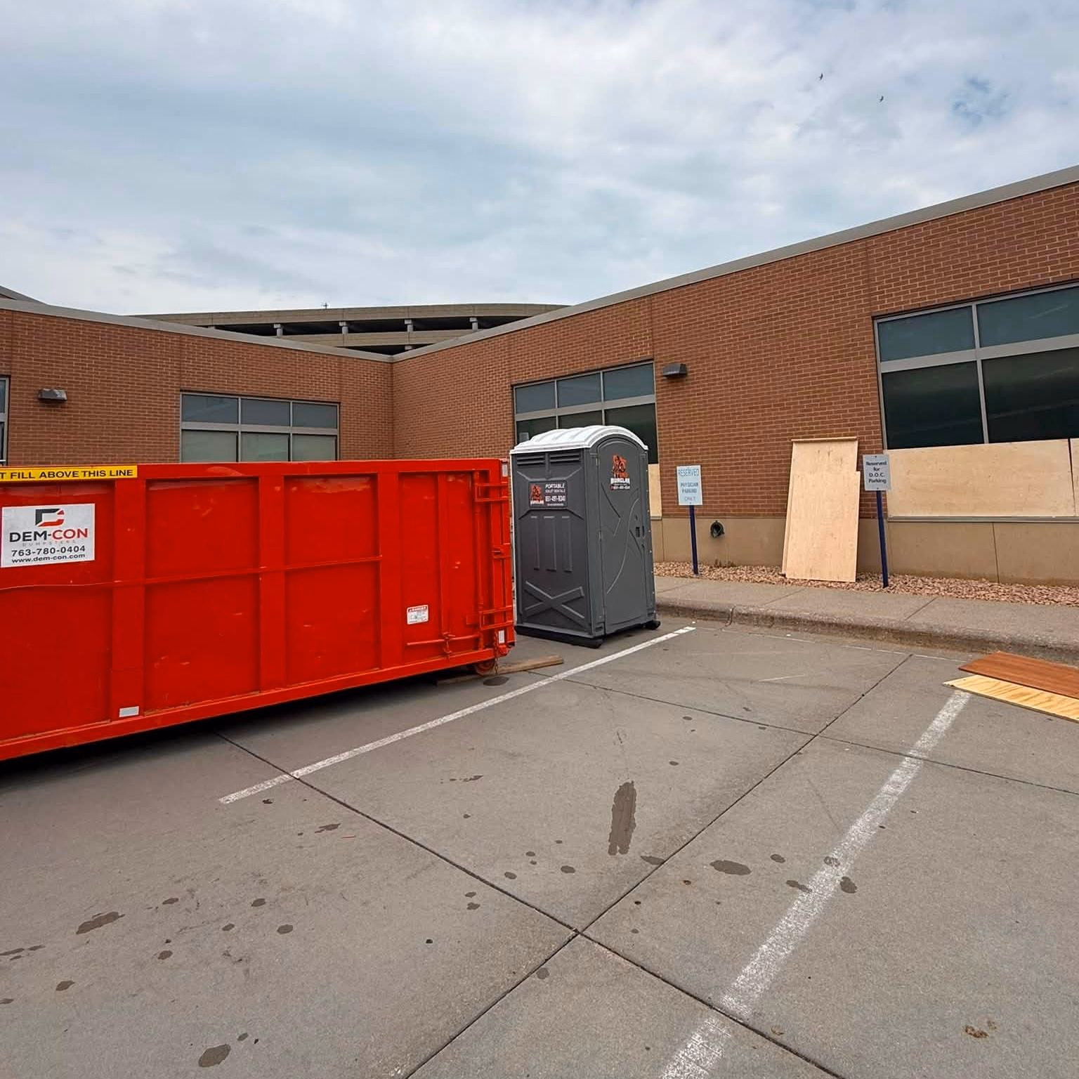 A portable toilet and a red waste container on the paved lot of a brick commercial building.