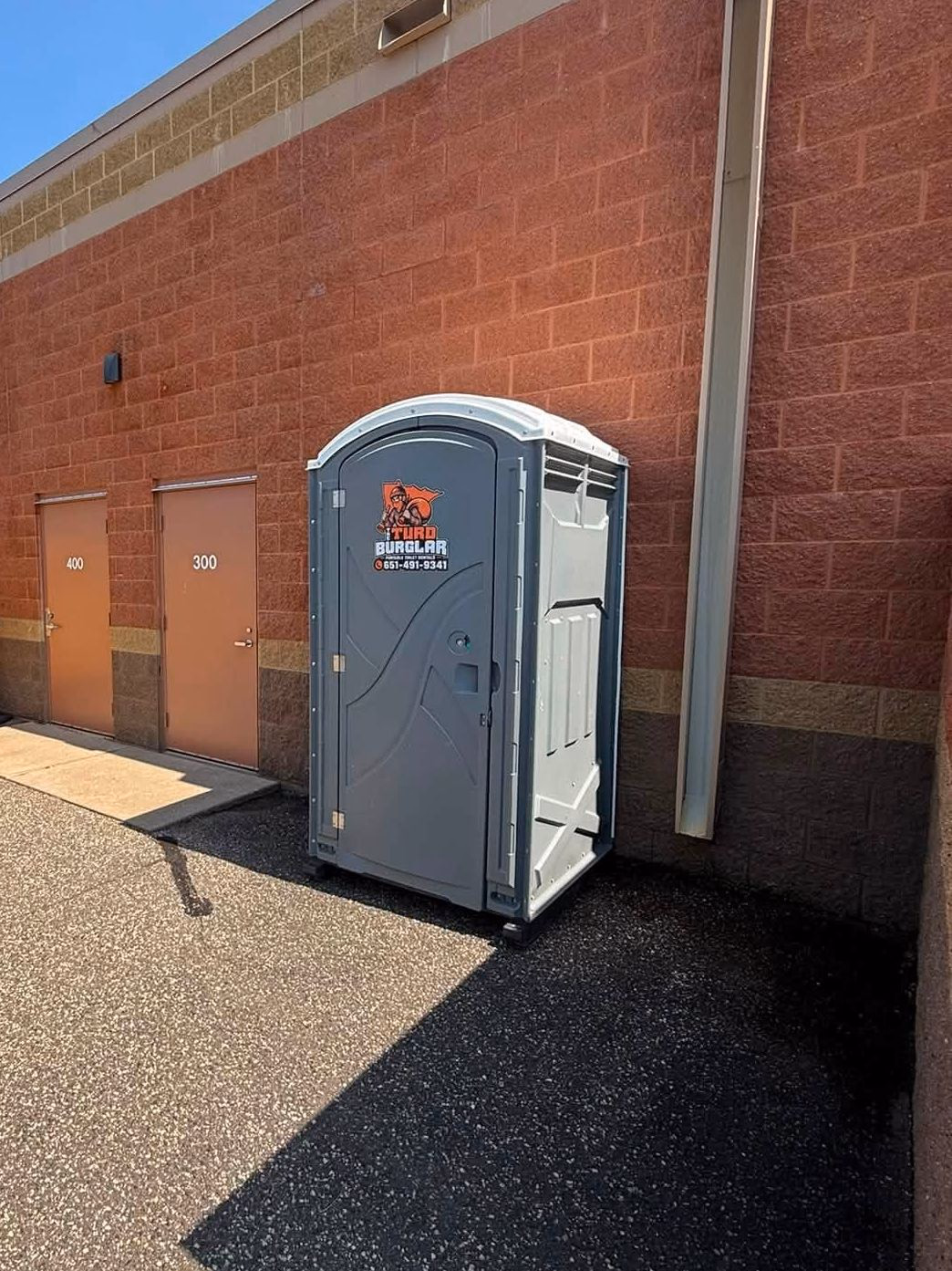 A gray portable toilet beside a yellow waste container against a brick commercial building.