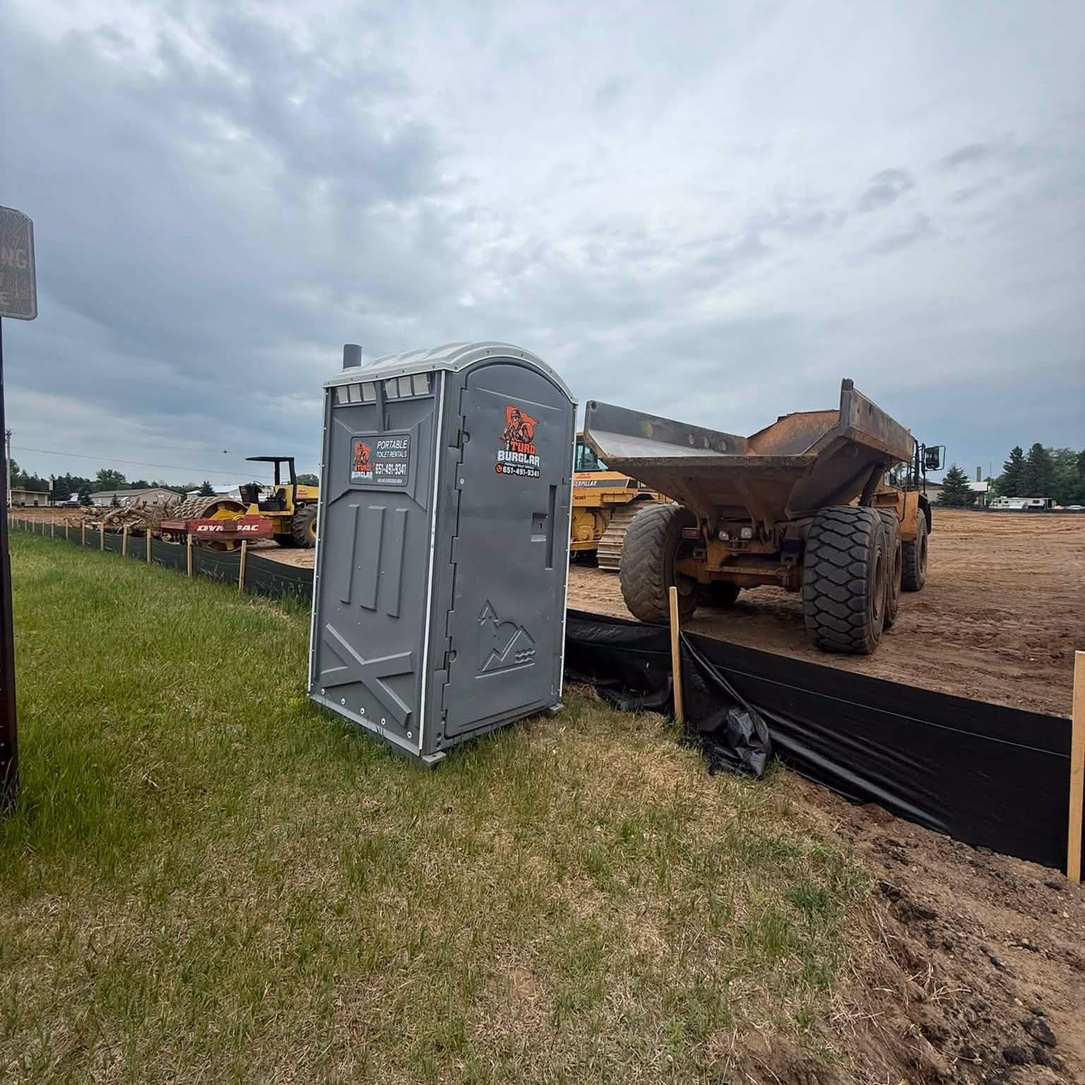 A gray portable toilet on a freshly graded lot next to an excavation trailer.