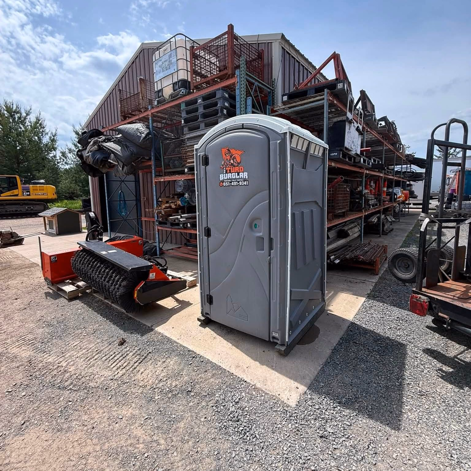 A gray portable toilet in an industrial yard surrounded by machinery and steel racking.