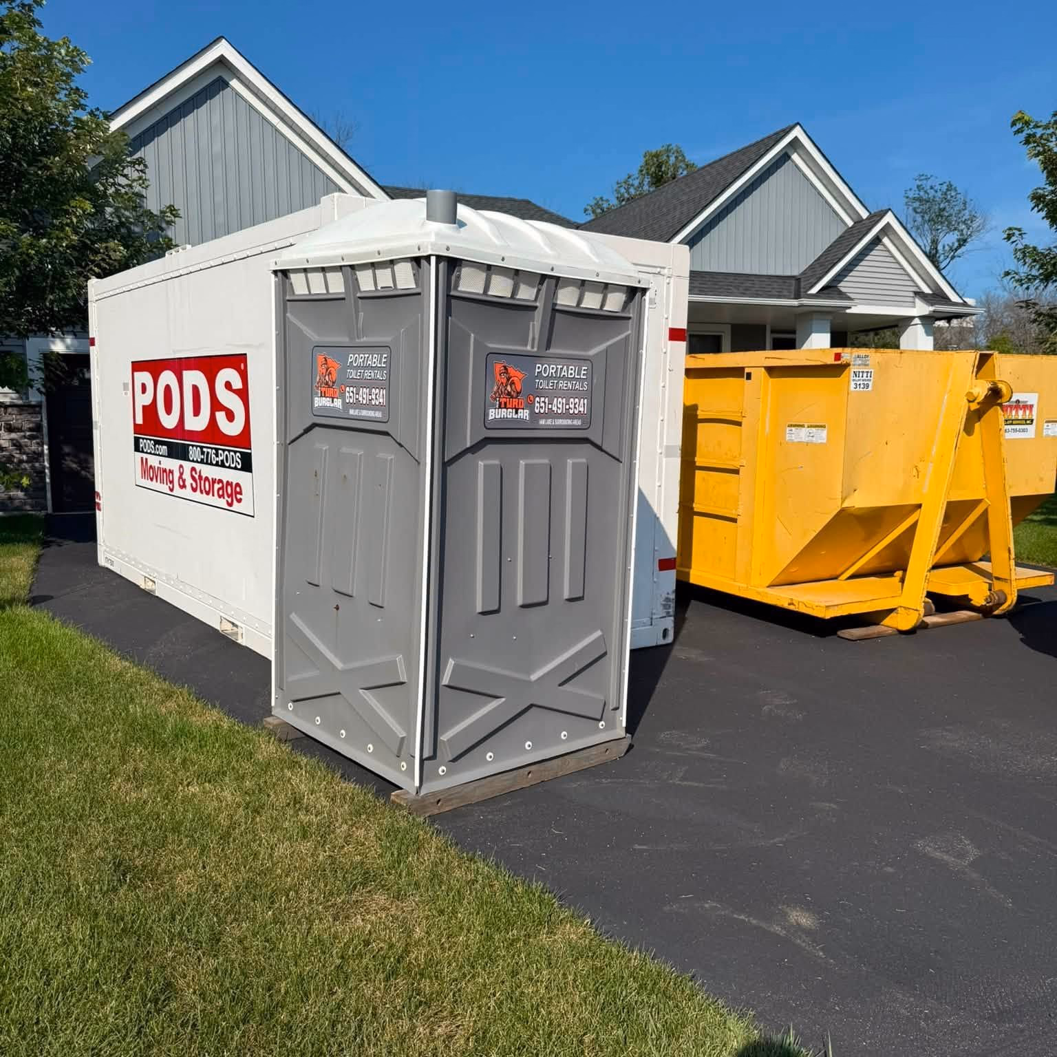 A portable toilet parked between a storage container and a yellow dumpster outside a new home.