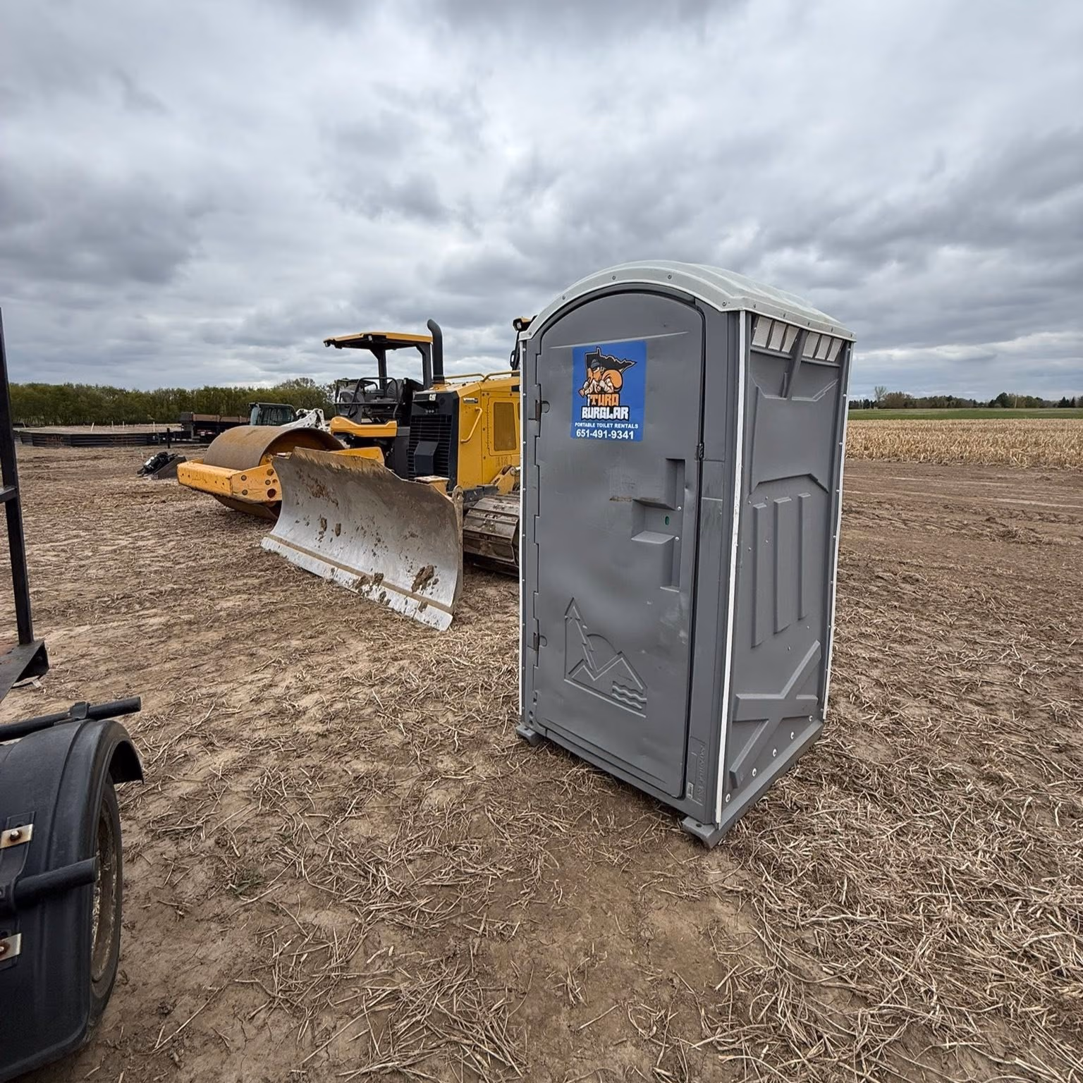 Gray portable toilet with a cartoon logo near a yellow bulldozer in an open dirt field under cloudy sky.