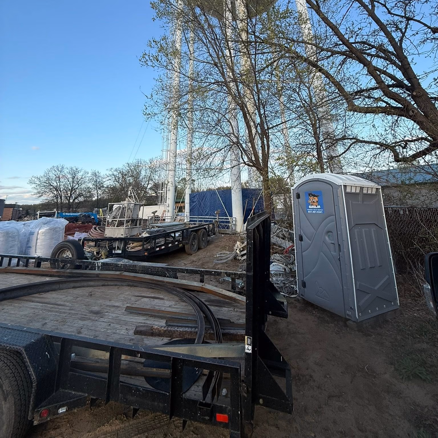 Portable gray porta potty at a construction site with trailers, equipment, and trees under blue sky.