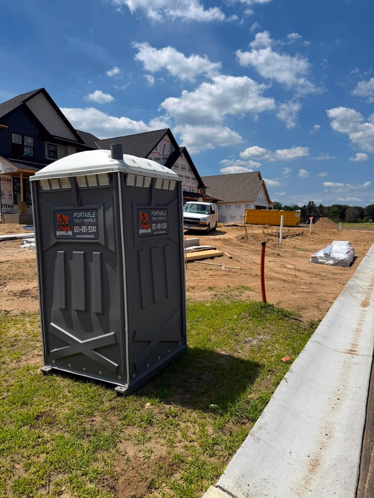 Gray portable toilet on grassy area at a housing construction site under blue sky with clouds.