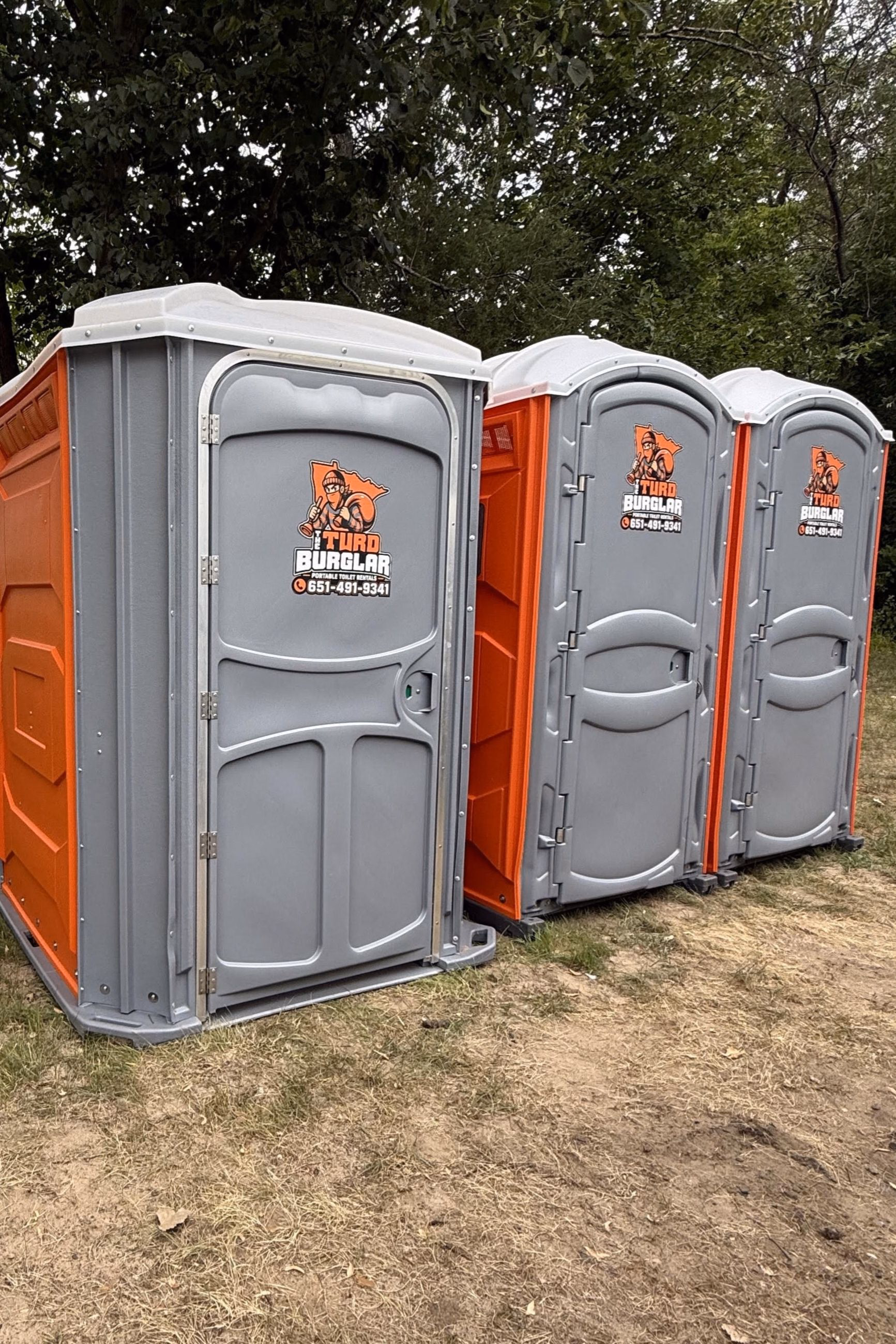 Three Turd Burglar portable toilets lined up on grass at the edge of a treeline.