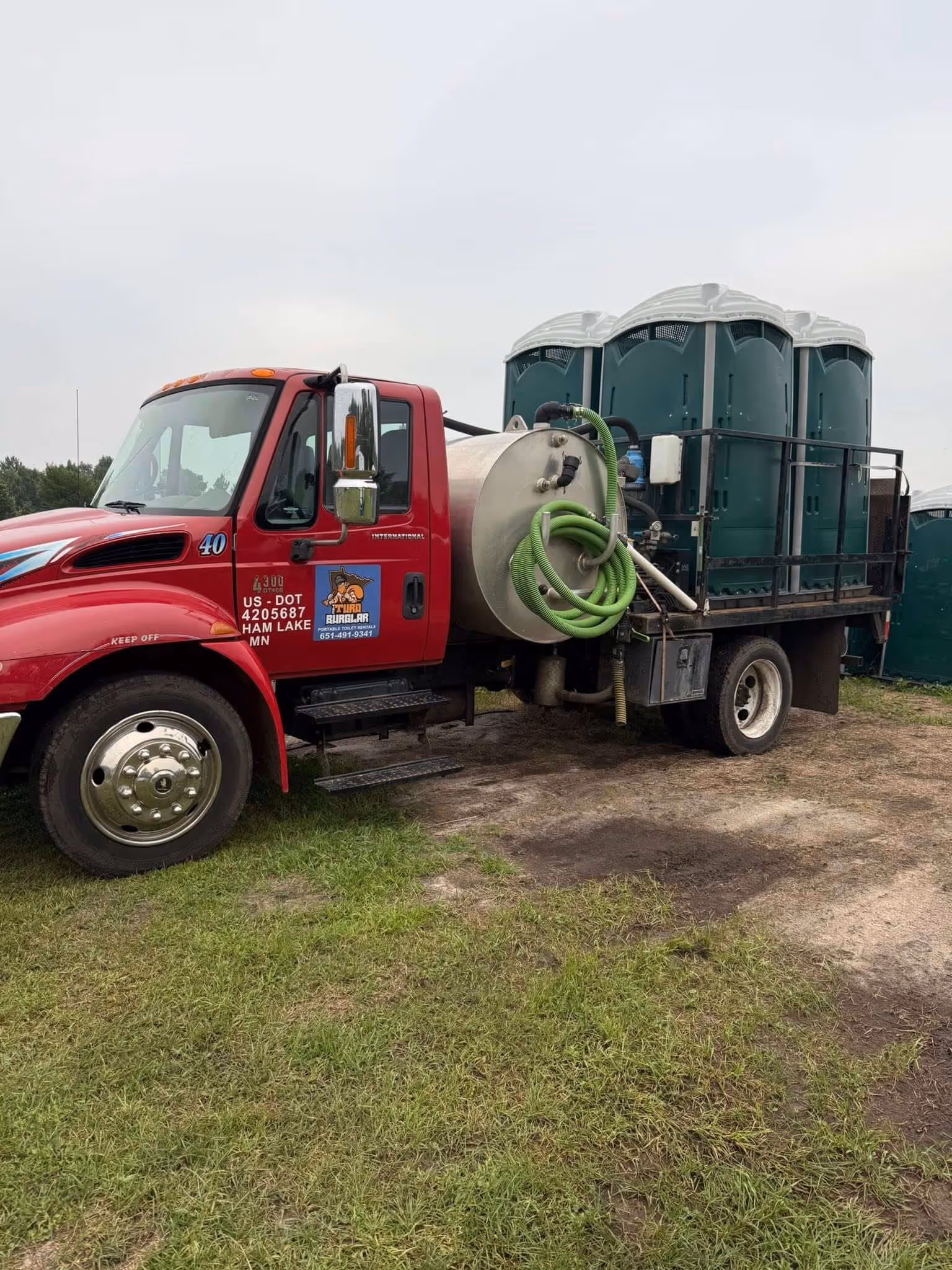 Red truck with large cylindrical tank and green hoses carrying portable toilets on a grassy area.