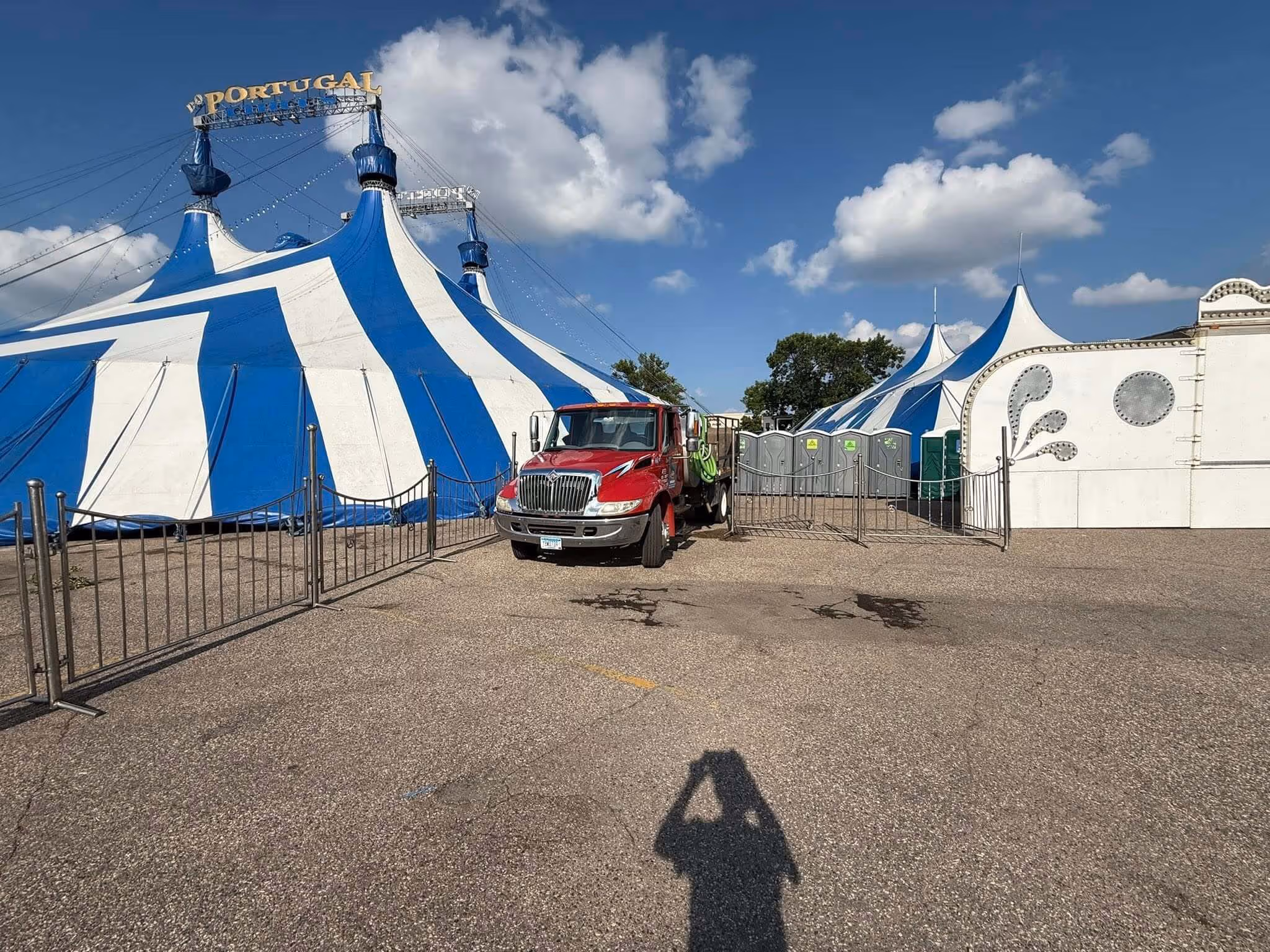 Large blue and white circus tents with a red truck parked in front and portable toilets visible behind a metal fence under a partly cloudy sky.