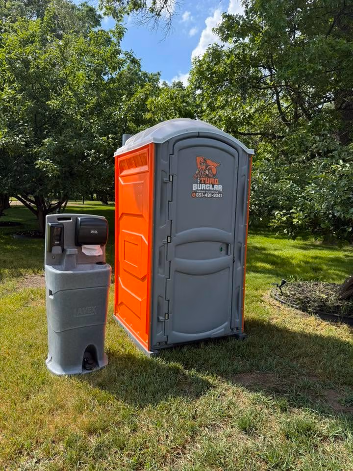 A row of seven green portable toilets with white roofs standing on wet pavement in a park-like area with trees in the background.
