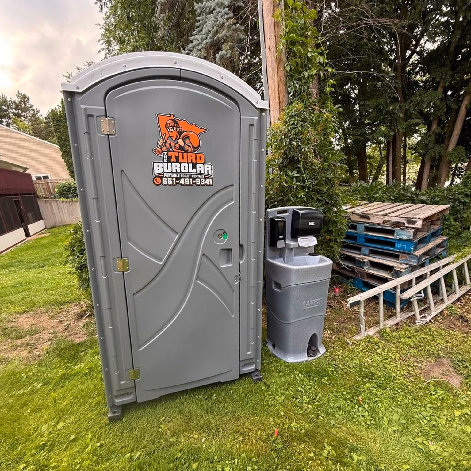 Gray portable toilet with a handwashing station and stacked pallets on grass beside trees.