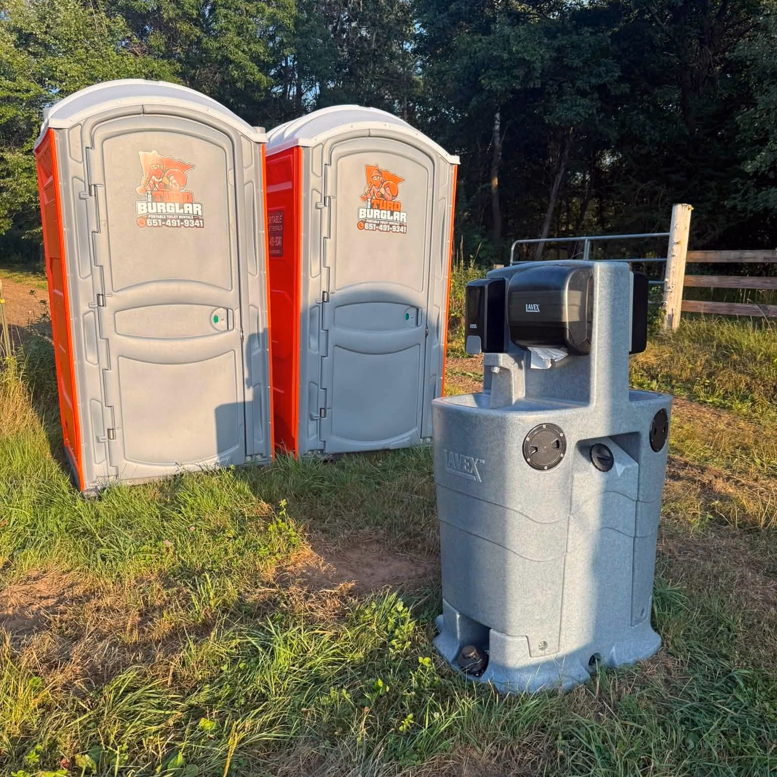 A portable toilet standing next to a gray hand wash station on an open lawn.