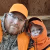 Man with beard and glasses wearing an orange cap and vest next to a child in an orange hooded jacket, both smiling indoors.