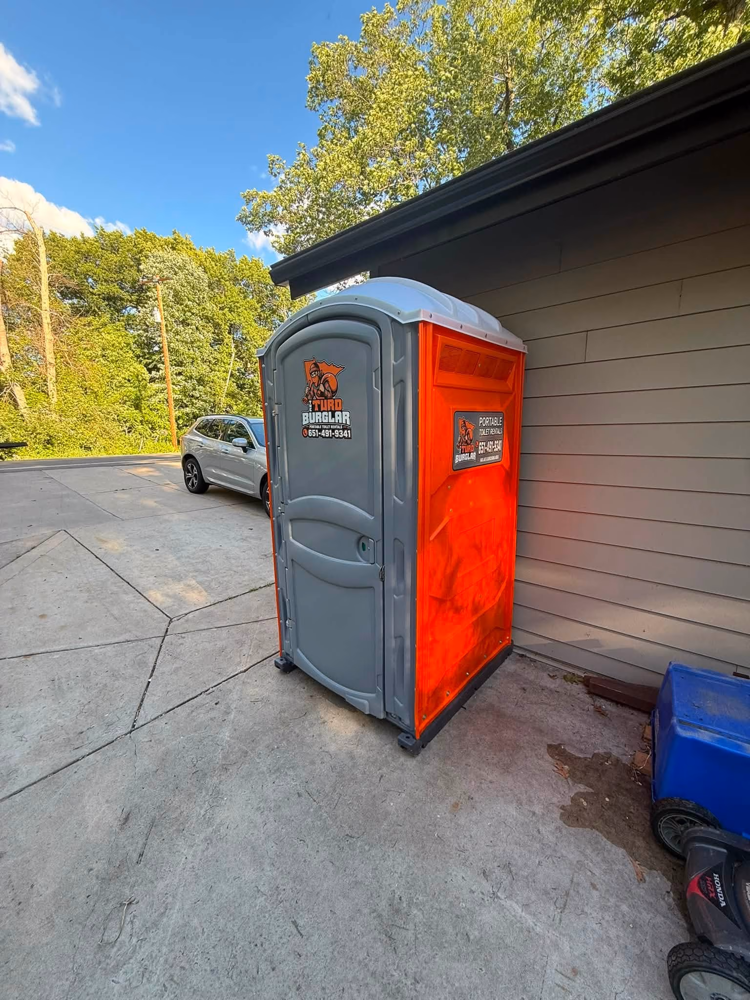 Orange and gray portable toilet with Turd Burglar signage on a concrete driveway near a building.