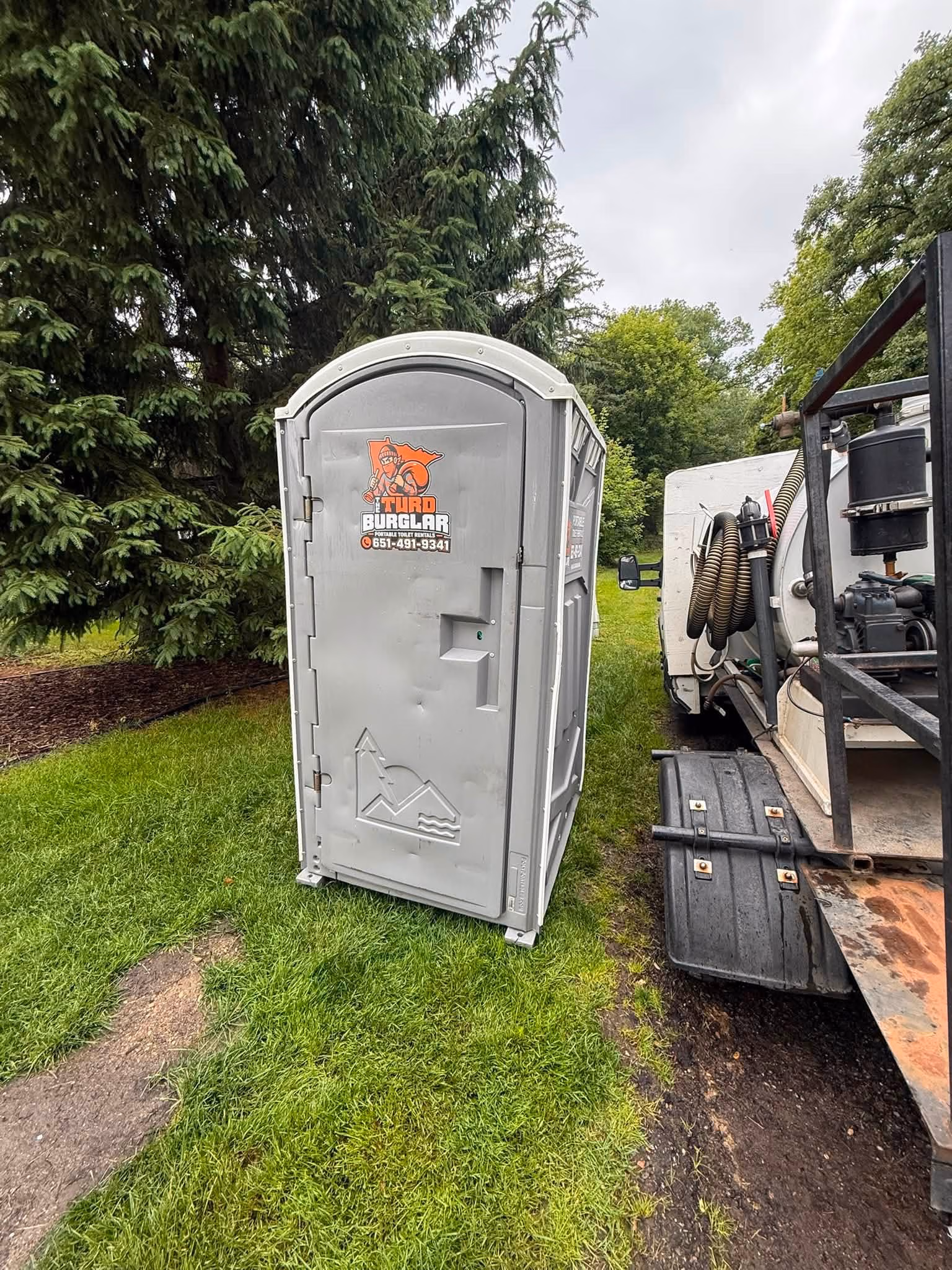 Row of green portable toilets mounted on a flatbed trailer in a grassy field under a partly cloudy sky.
