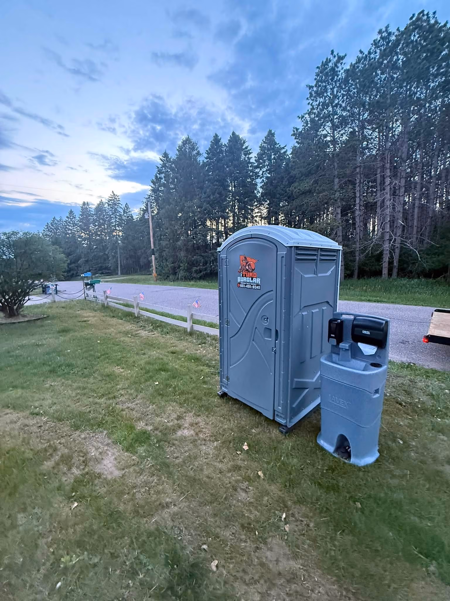 Red sanitation truck carrying green portable toilets on a grassy area under a cloudy sky.