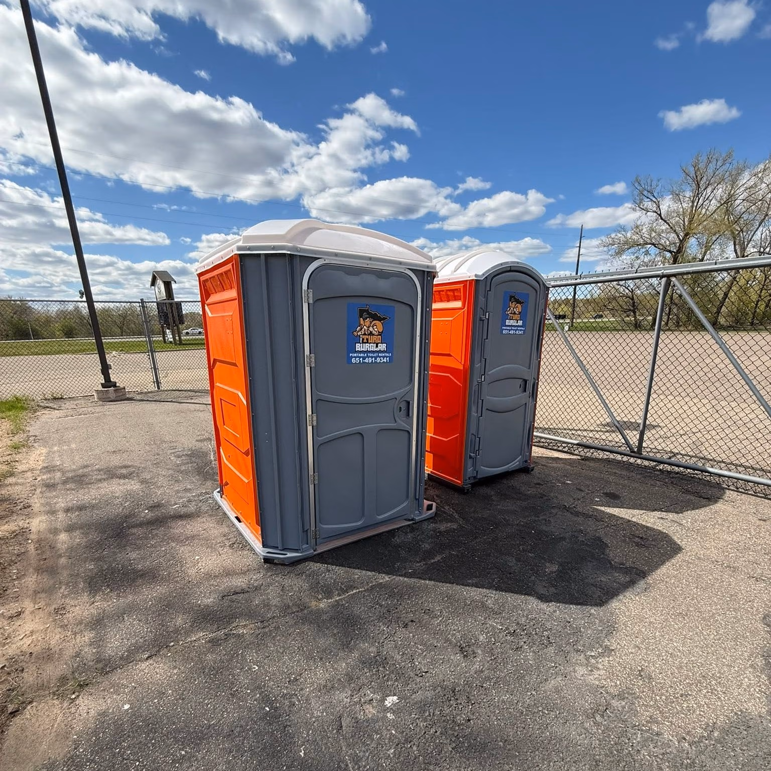 Two orange and gray portable toilets standing on pavement near a chain-link fence under a blue sky.