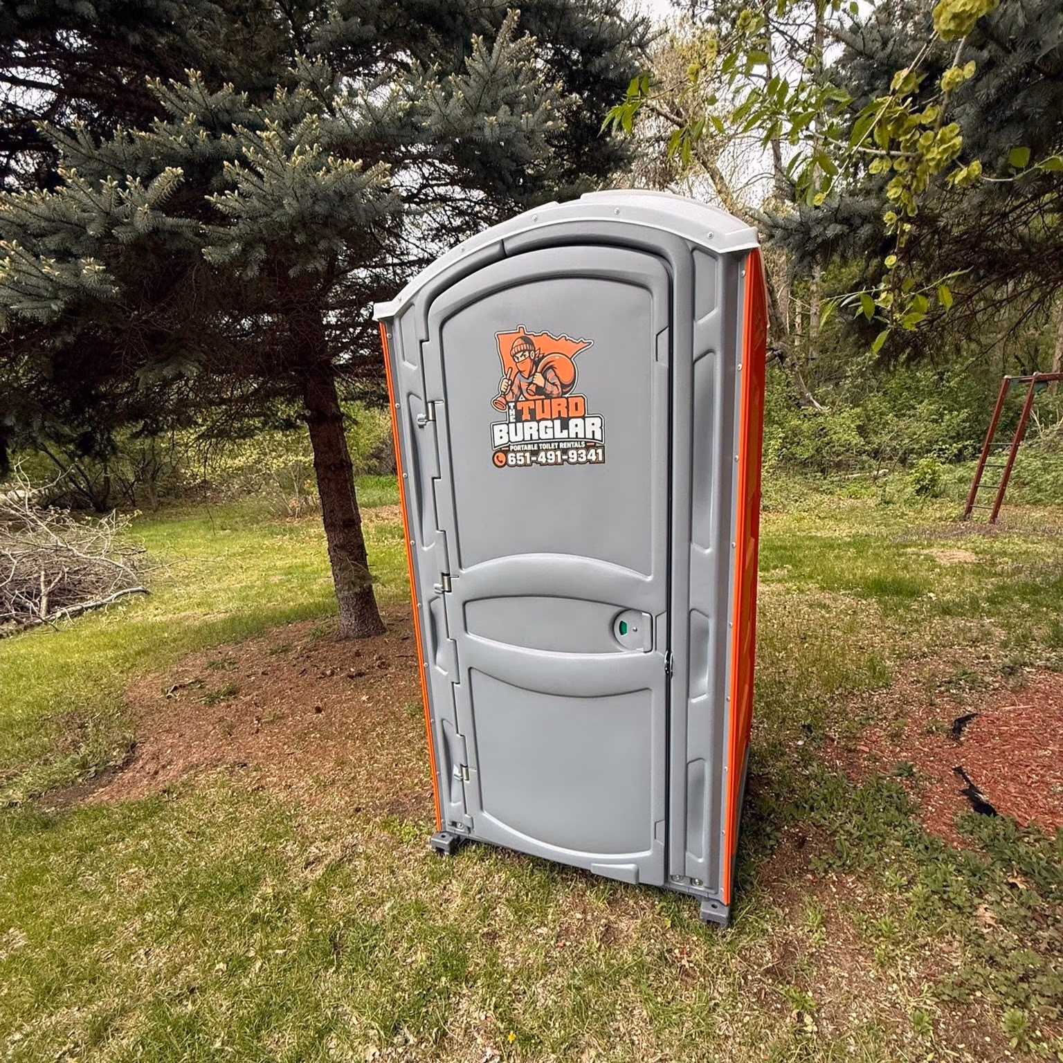 Gray portable toilet with 'Turd Burglar' logo and contact number, placed against a brick building under a cloudy sky.