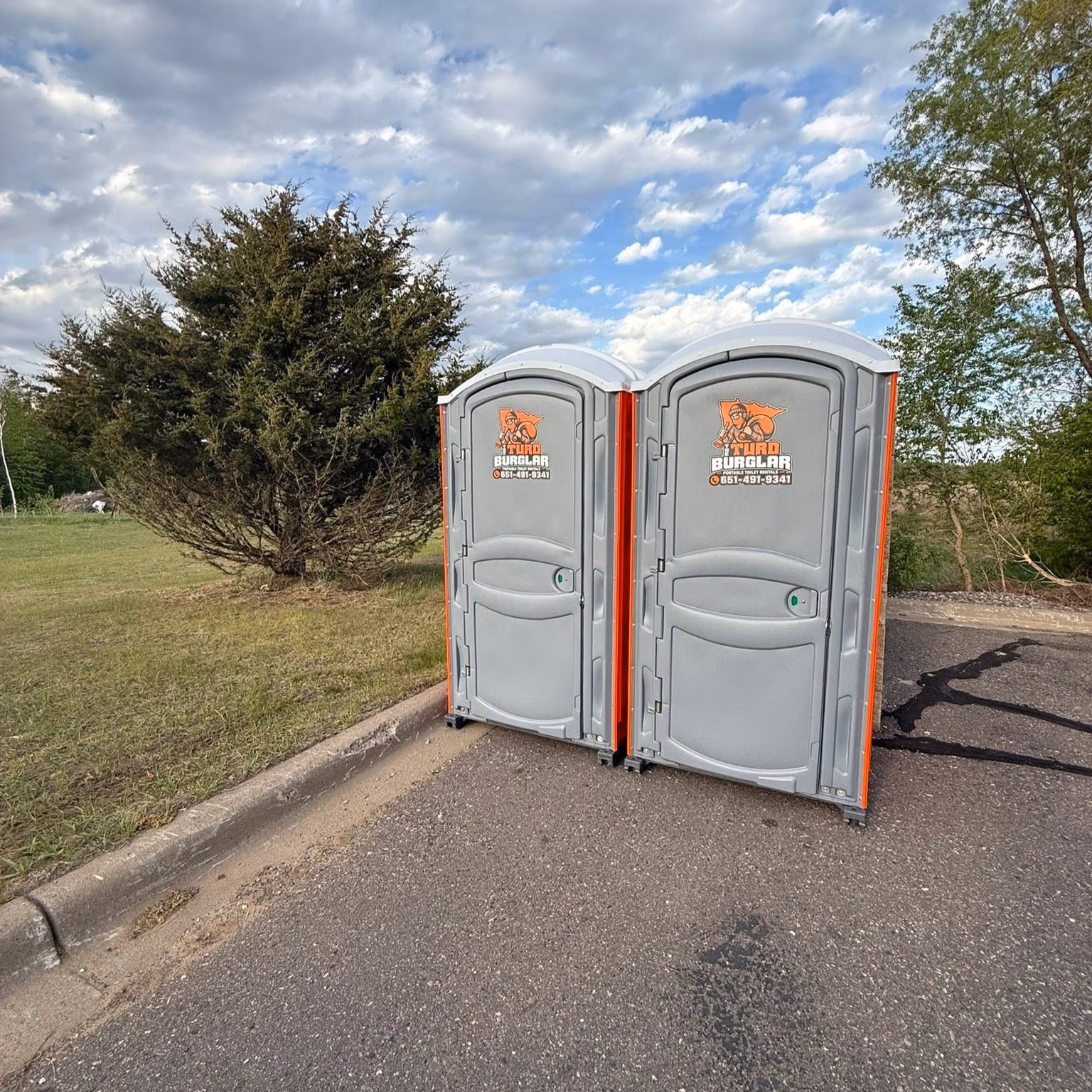 Alley beside a building with a grey portable toilet labeled Turbo Burglar and a white service truck with coiled yellow and black hoses.