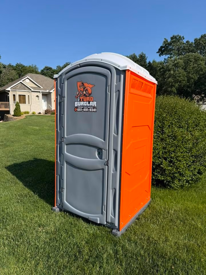 Orange and gray portable toilet on green grass with a house and bushes in the background.