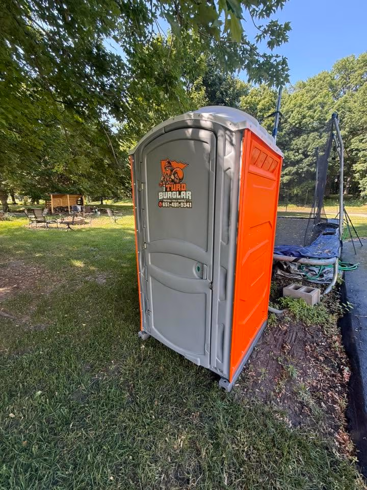 Gray and orange portable toilet with 'Turd Burglar' logo on grass near a trampoline and trees.