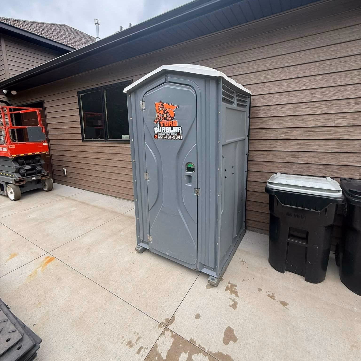 A gray portable toilet outside a house with a scissor lift parked on the driveway.