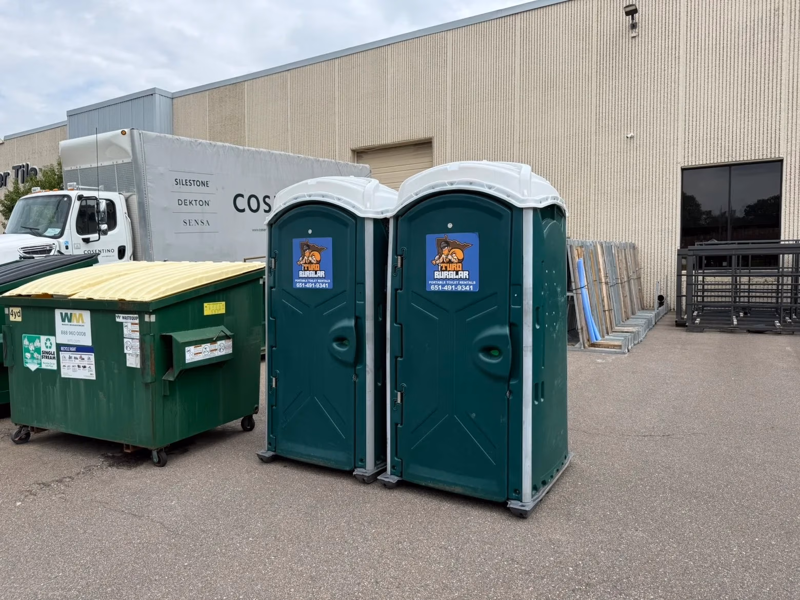Two green portable toilets labeled 'Turbo Burglar Portable Toilet Rentals' next to a green and yellow dumpster and a white truck parked by a beige warehouse building.