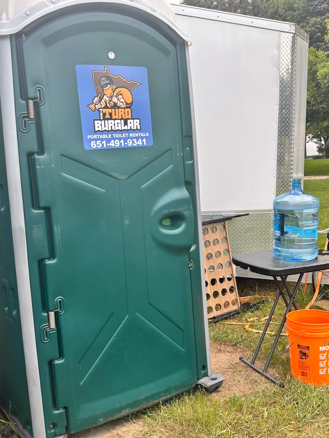 Green portable toilet with The Turd Burglar logo and phone number, near a table with a large water jug and orange bucket.