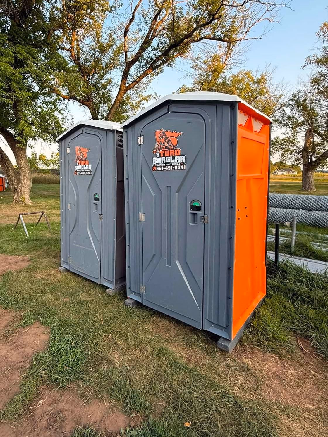 Two portable toilets on a bare dirt lot with trees and outbuildings behind them.