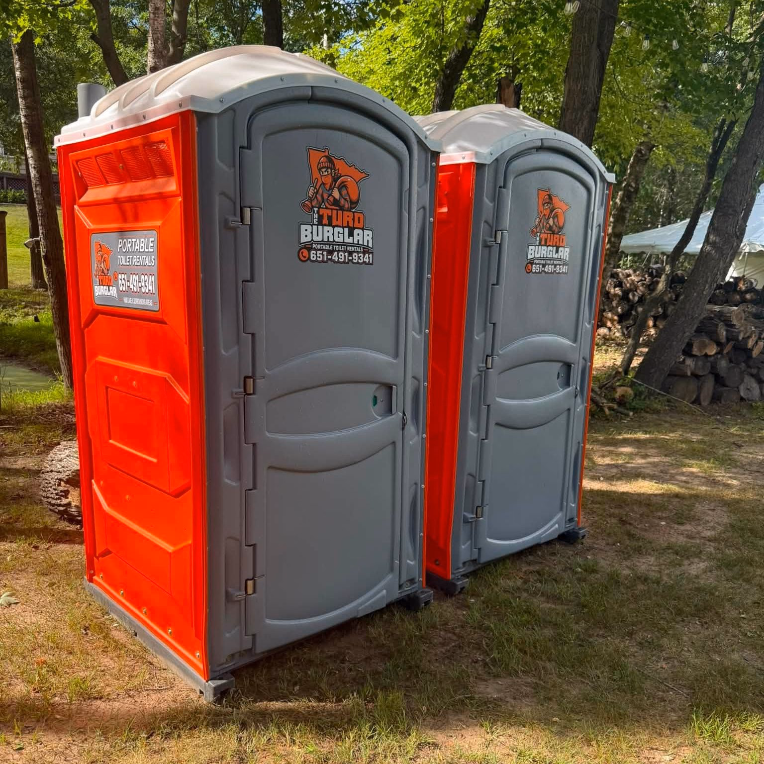 Two portable toilets standing together on grass at the edge of a tree line.