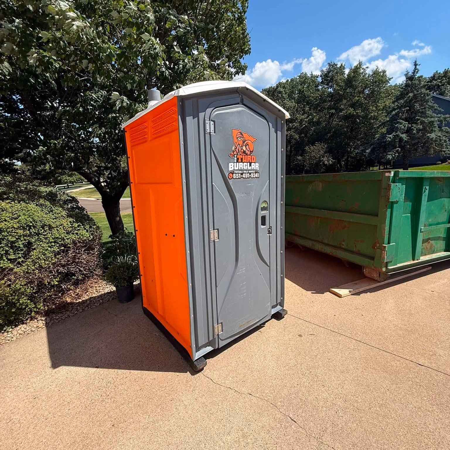 An orange and gray portable toilet standing beside a green roll-off dumpster on a paved drive.