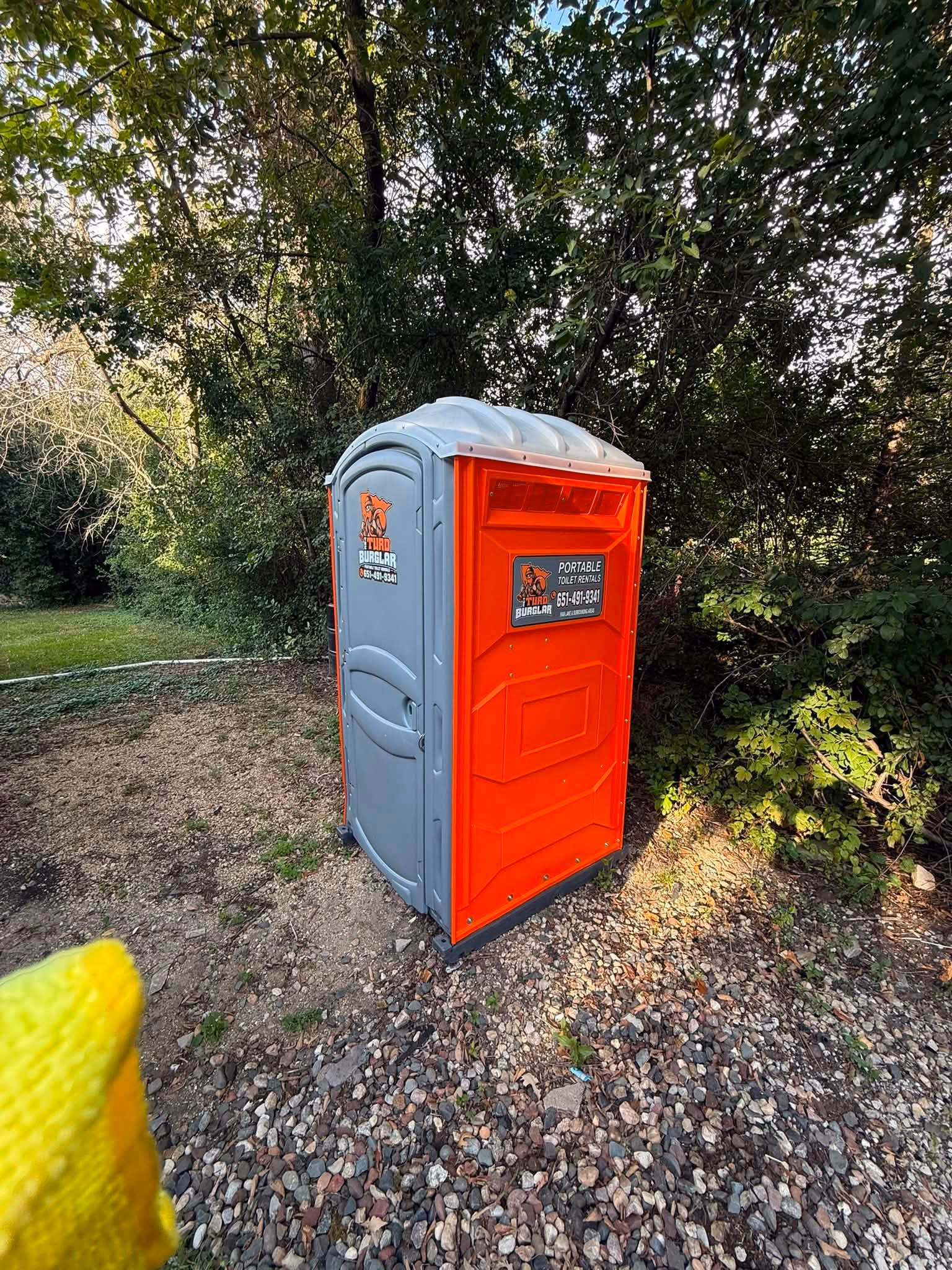 An orange and gray portable toilet set on a gravel pad among trees.