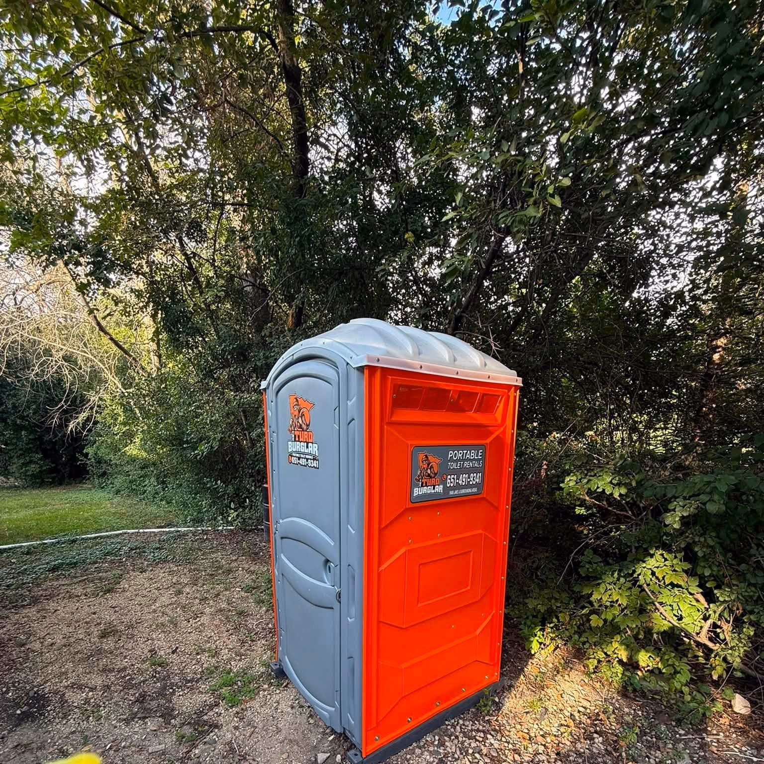 An orange and gray portable toilet set on a gravel pad among trees.