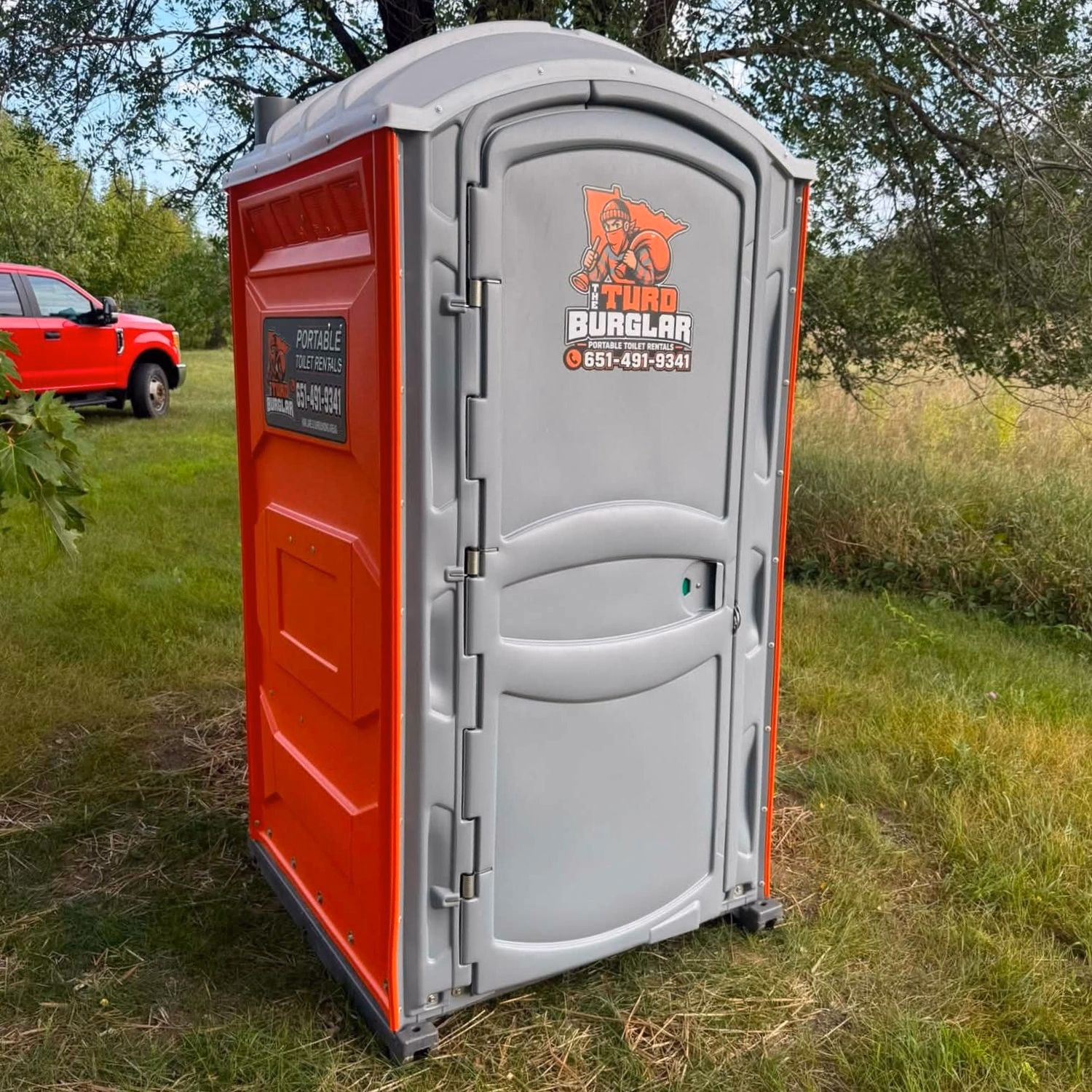 An orange and gray portable toilet on a mown lawn with a red pickup parked nearby.