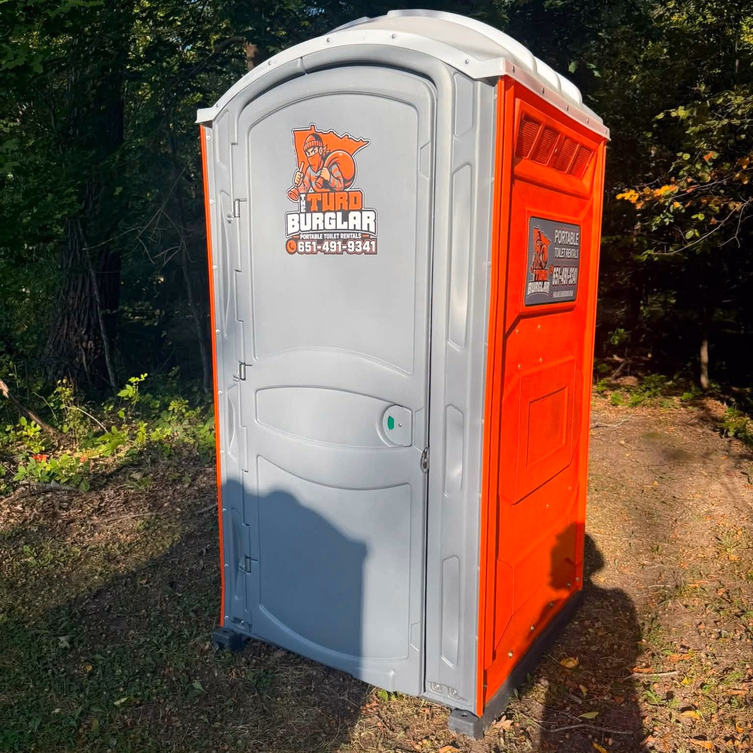 An orange and gray portable toilet on a wooded lot in low evening light.