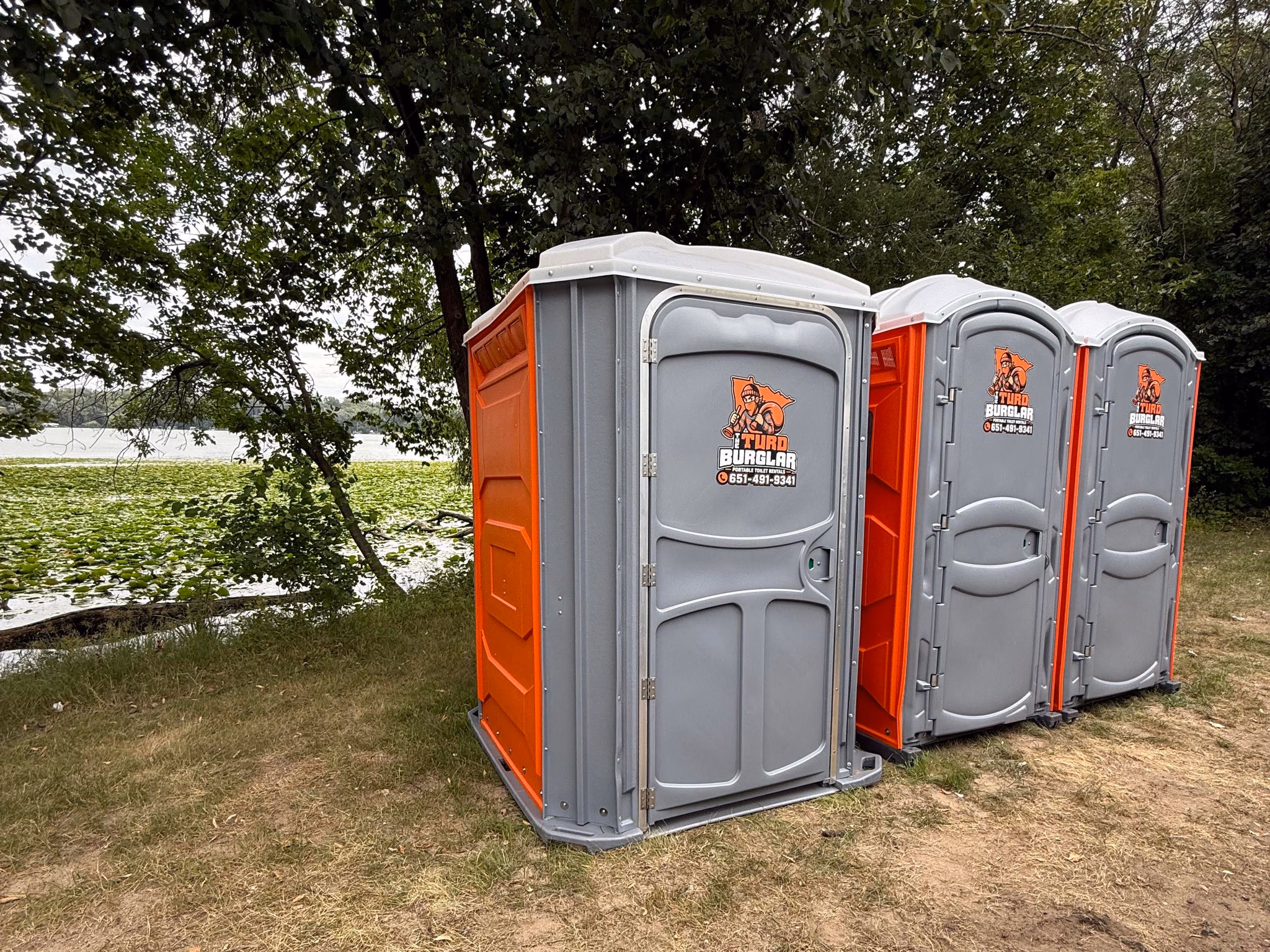 Three orange and gray portable toilets standing in a row on grass beside a lake.
